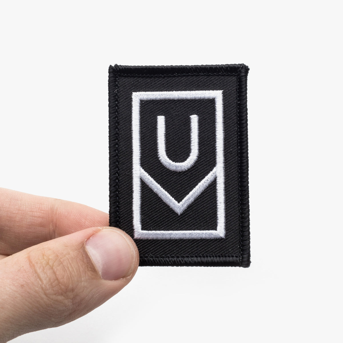 A hand holds the Ugmonk Patch, a rectangular black patch featuring bold white geometric lines that form abstract shapes—possibly suggesting the letters U and V—against a plain white background.