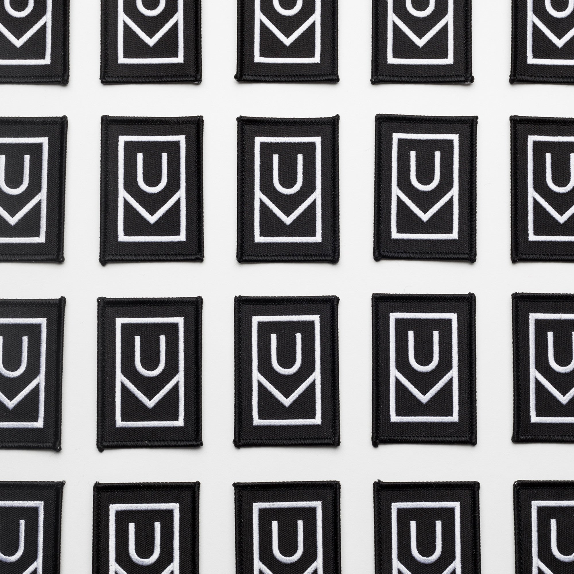 Multiple Ugmonk Patches—black rectangular embroidered patches featuring a bold white geometric U-shaped design—are arranged in neat rows on a white background.