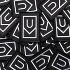 A pile of Ugmonk Patches—black, rectangular embroidered patches featuring white geometric designs with a U shape above a downward-pointing arrow.