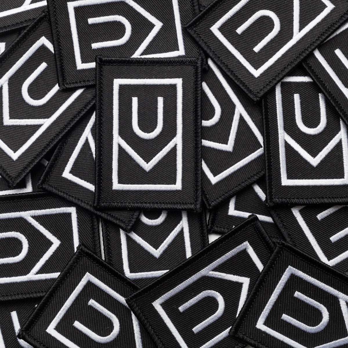 A pile of Ugmonk Patches—black, rectangular embroidered patches featuring white geometric designs with a U shape above a downward-pointing arrow.
