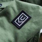 A close-up of an olive green fabric with a black rectangular Ugmonk Patch, featuring a white geometric logo made up of the letters C and U stitched onto it.