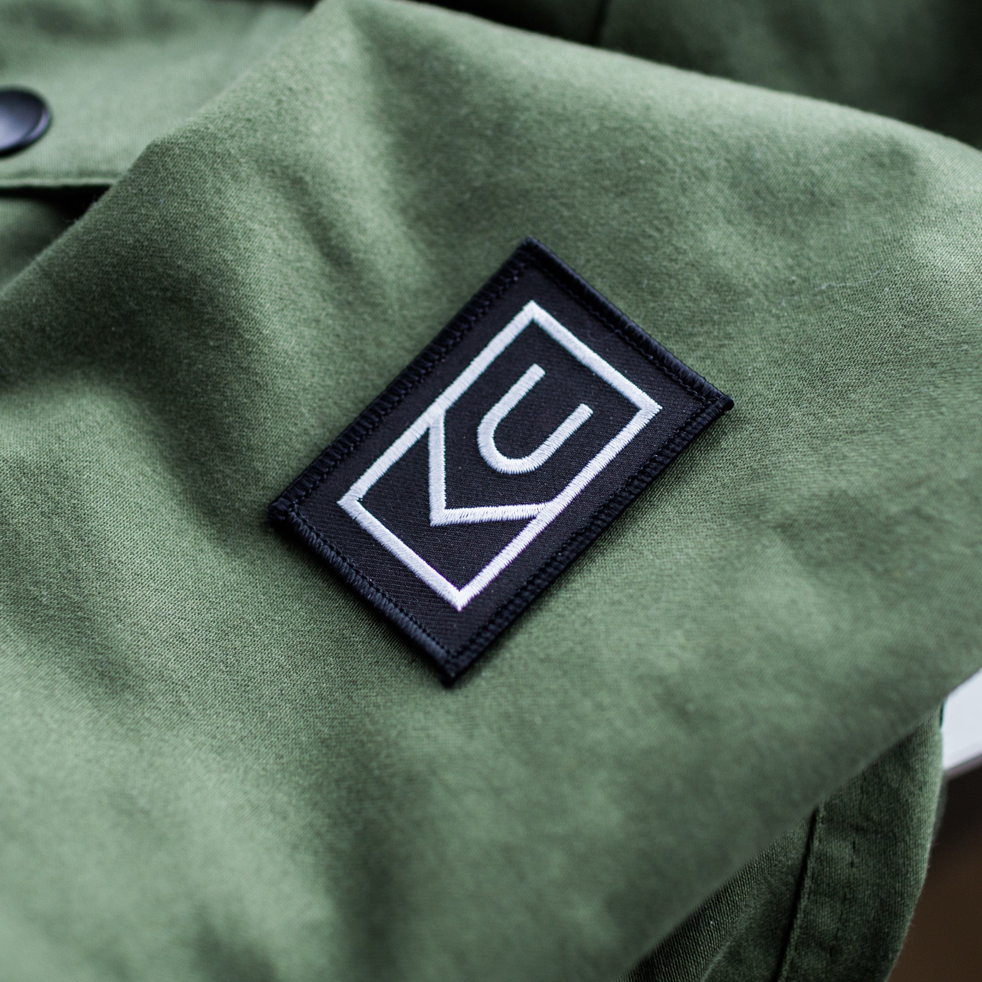 A close-up of an olive green fabric with a black rectangular Ugmonk Patch, featuring a white geometric logo made up of the letters C and U stitched onto it.