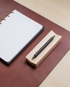 A dotted notebook with gold rings lies open on a brown desk pad, next to a black pen resting on the Pen Tray (Maple), all on a light wood desk. The Pen Tray (Maple) is expertly made in Pennsylvania.