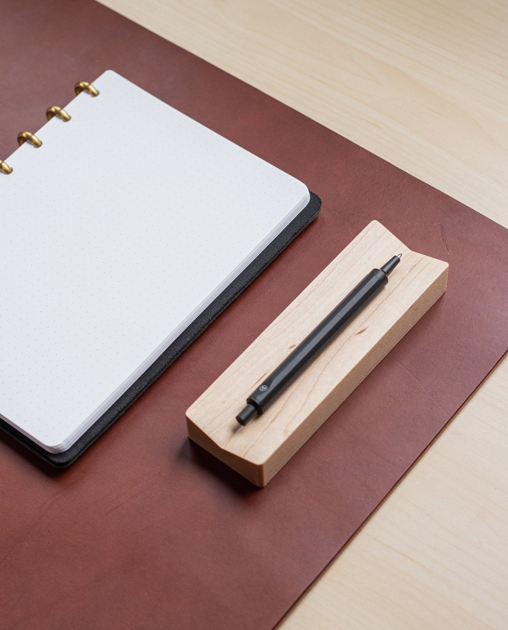 A dotted notebook with gold rings lies open on a brown desk pad, next to a black pen resting on the Pen Tray (Maple), all on a light wood desk. The Pen Tray (Maple) is expertly made in Pennsylvania.