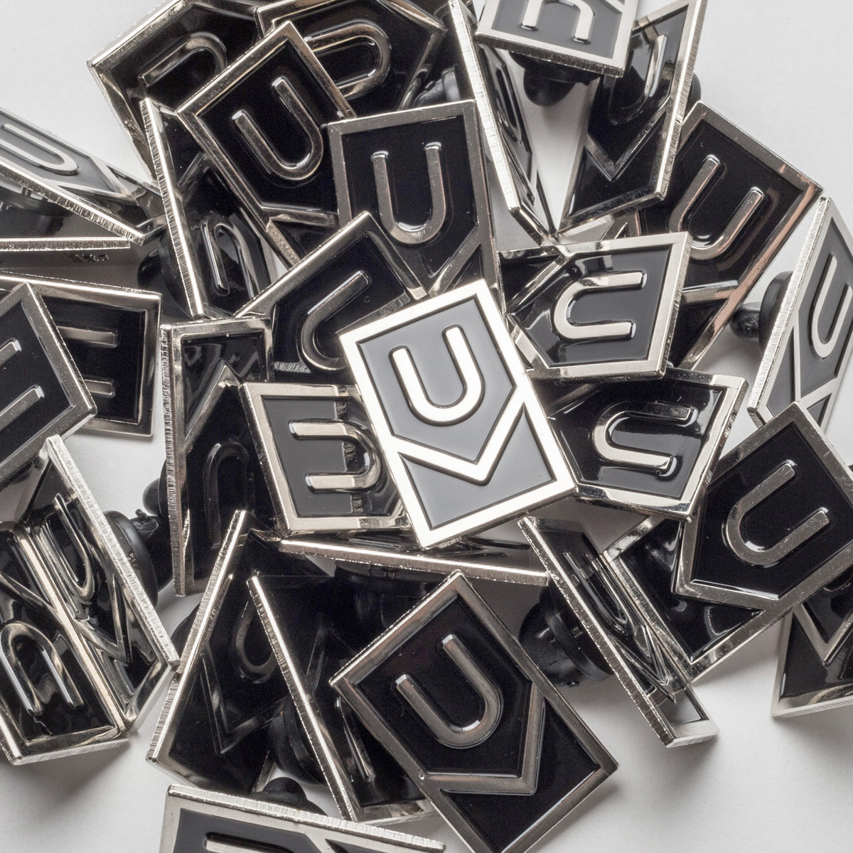 A collection of Ugmonk Enamel Pins with a geometric U-shaped design in black and silver, scattered and overlapping on a white surface.