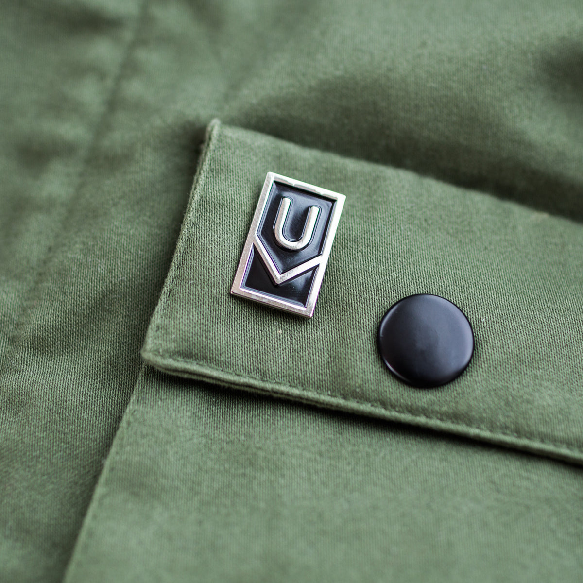 A close-up of olive green fabric featuring a black snap button and the Ugmonk Enamel Pin—a rectangular silver logo with a geometric design displaying the letters U and V.