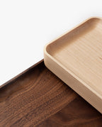 Two rectangular wooden trays overlap on a white background; one handcrafted valet tray (Maple – Small) features light-colored solid maple with rounded corners, while the other tray is darker with visible wood grain.