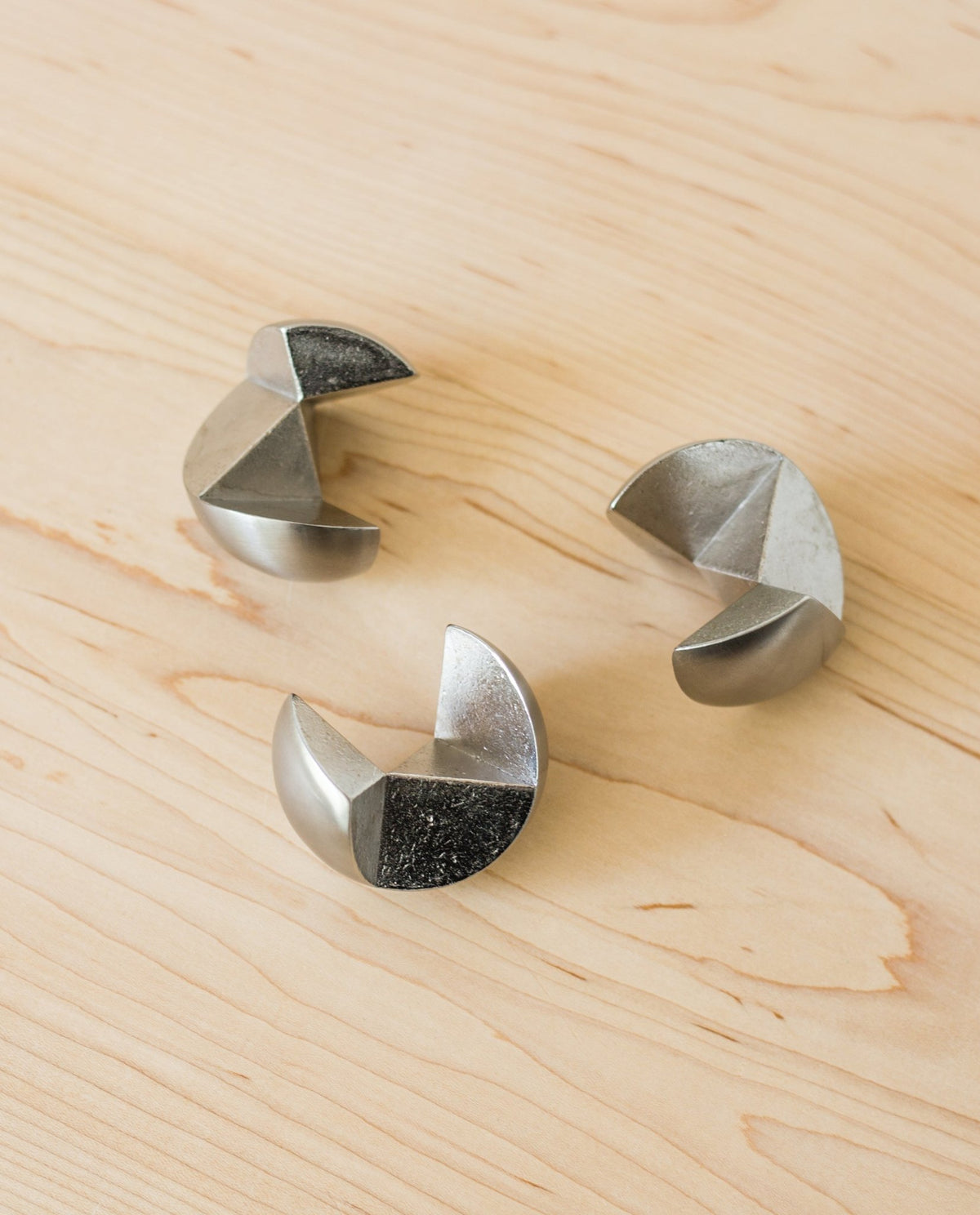Three stainless steel geometric pieces from the Craighill Venn Puzzle, featuring crescent and triangular shapes, are arranged in a loose triangle on a light wood surface.