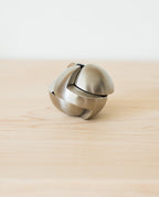 The Craighill Venn Puzzle (Stainless Steel) is displayed assembled on a light wooden surface, its smooth, curved pieces interlocking to form a striking spherical shape reminiscent of this renowned puzzle.