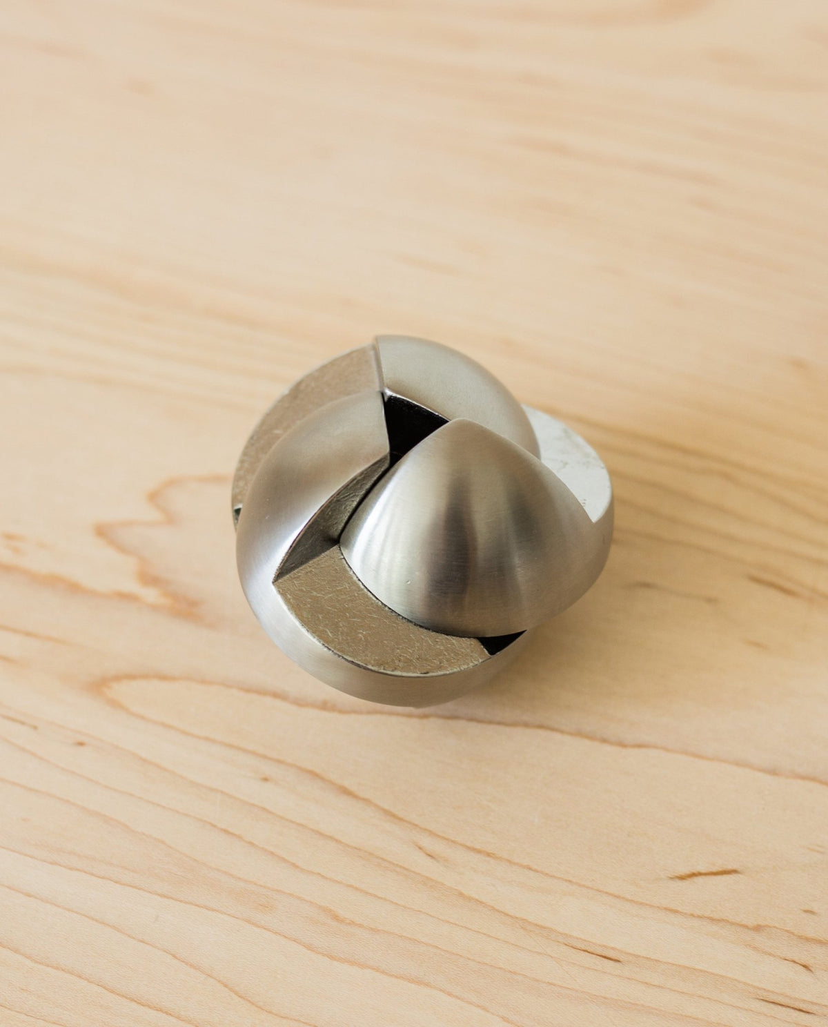 The Craighill Venn Puzzle (Stainless Steel) with interlocking, curved pieces rests on a light wood surface. This geometric, spherical mechanical puzzle has a smooth, shiny finish and is crafted from stainless steel.