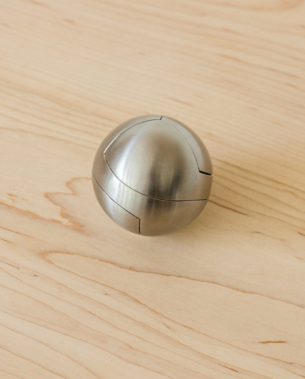 The Craighill Venn Puzzle (Stainless Steel) is a metallic, spherical puzzle with smooth, brushed surfaces and visible interlocking seams, resting on a light wooden surface.