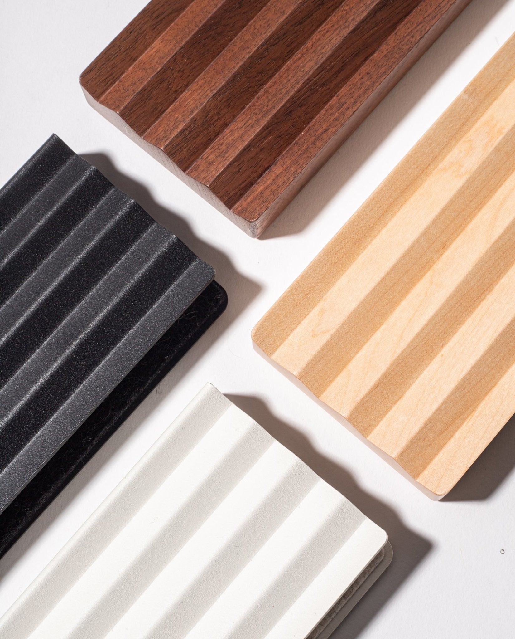 Four rectangular Gather Wood Zigzag Trays (2x1 - Walnut) with ribbed textures and varied colors are arranged diagonally on a white surface, their shadows adding dimension and highlighting the solid walnut option.
