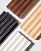 Four rectangular panels—one being the Gather Wood Zigzag Tray (2x1 - Maple) in solid maple and others in black, dark brown, light beige, and white finishes—each featuring parallel ridges characteristic of the tray's design, are arranged diagonally on a light surface with their corners converging toward the center of the image.